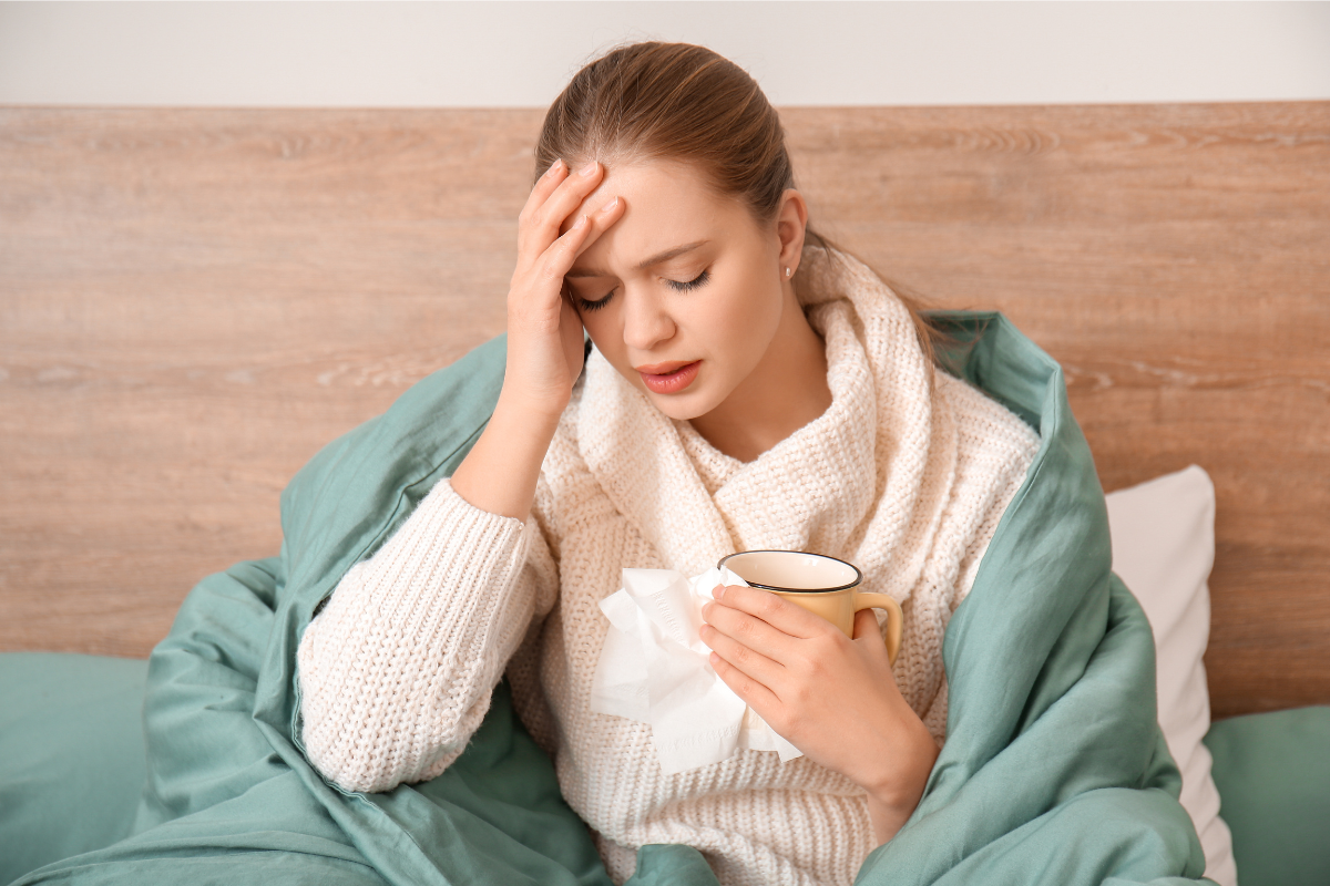 Woman sitting in bed, wrapped in a blanket, holding a cup and touching her forehead, showing signs of fatigue and discomfort.