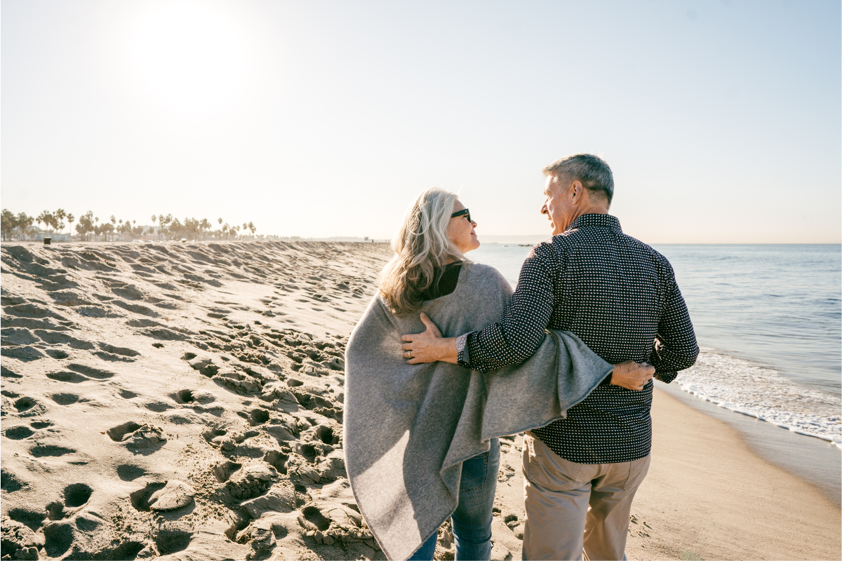 A senior couple walking arm in arm on the beach, enjoying a sunny day.