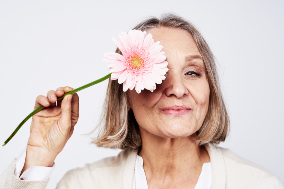 A joyful older woman with shoulder-length blonde hair holds a pink daisy up to her eye, smiling warmly.