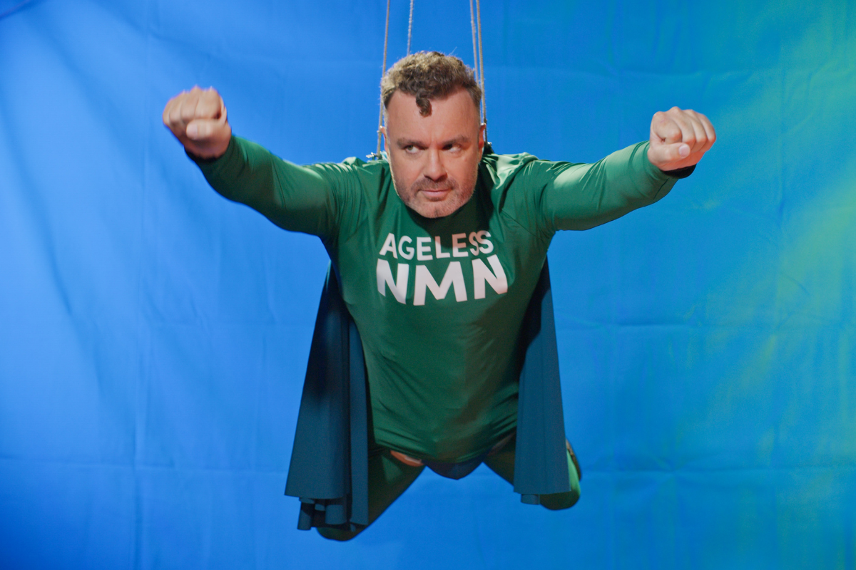 Man in green “Ageless NMN” superhero costume flying against blue background.