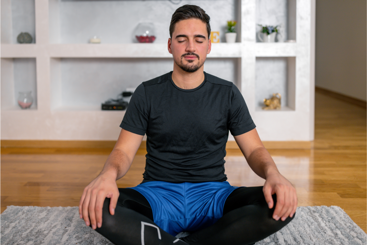 A man practising the Wim Hof breathing technique, seated comfortably on a rug in a modern living room, with eyes closed and a calm expression.