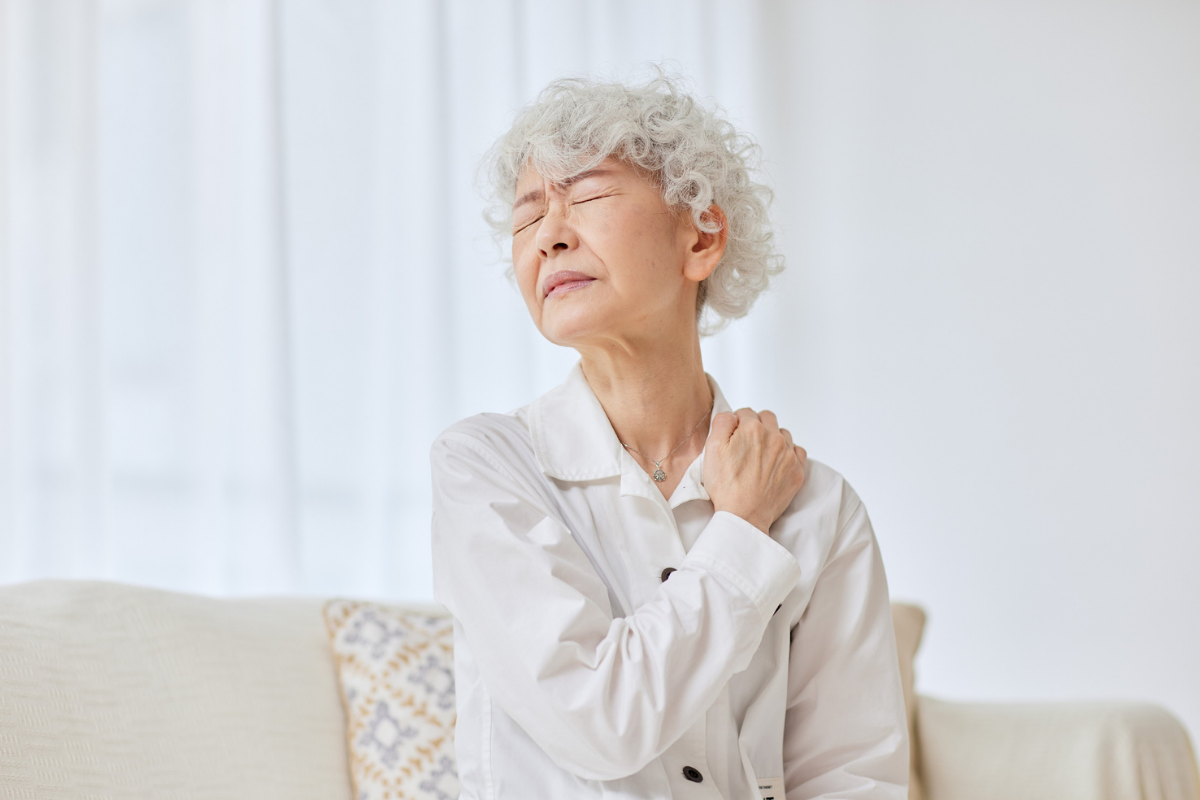 Older woman sitting on a sofa, touching her shoulder and wincing in discomfort.
