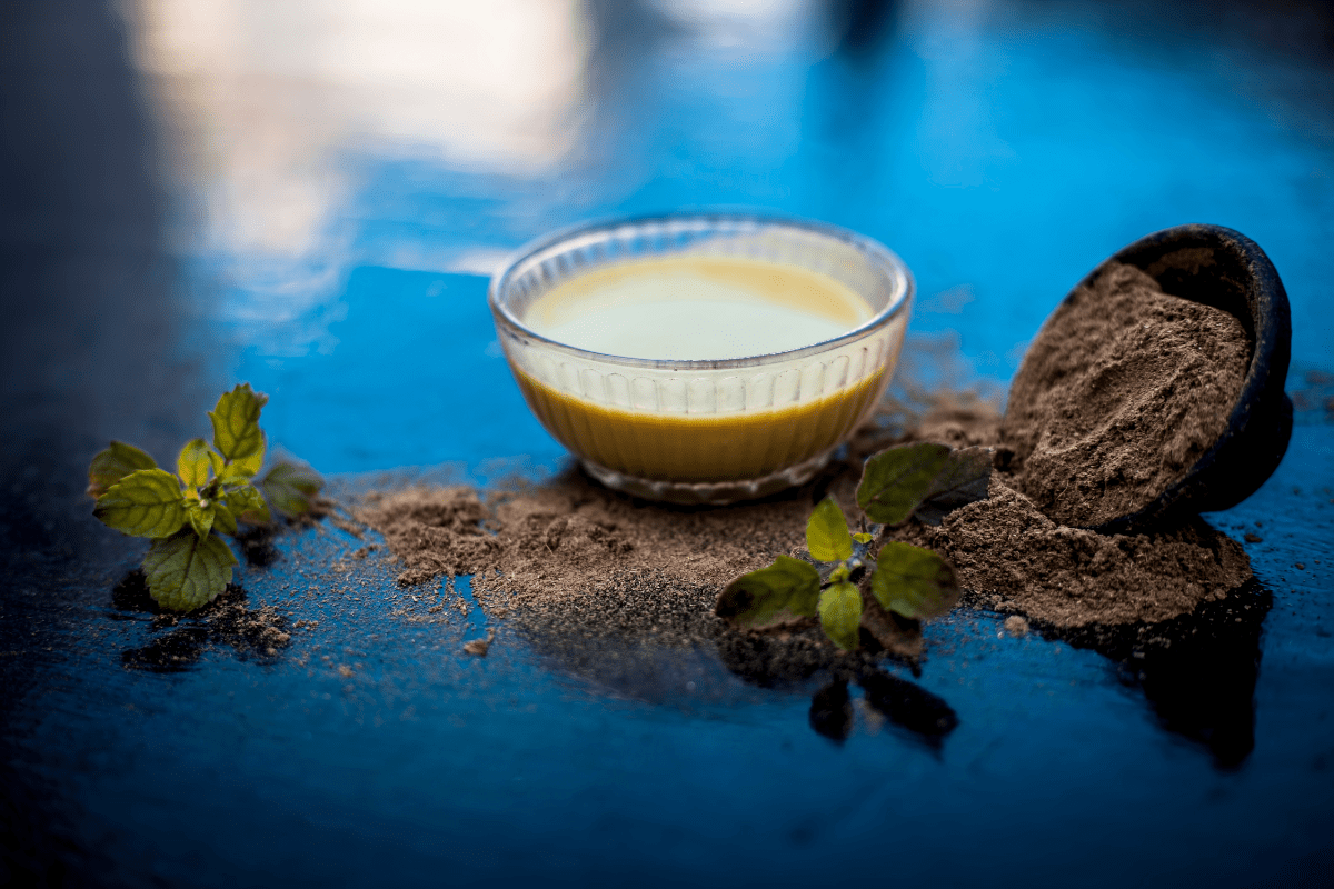 A bowl of herbal liquid surrounded by powdered herbs and fresh leaves on a reflective surface.