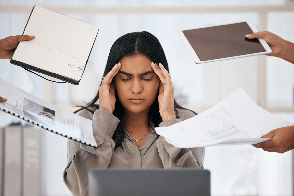 Stressed woman holding her head while surrounded by paperwork, devices, and tasks.