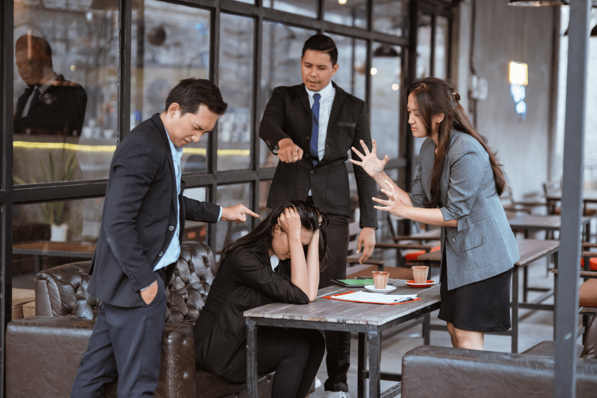 Stressed woman at a table surrounded by colleagues pointing and arguing.