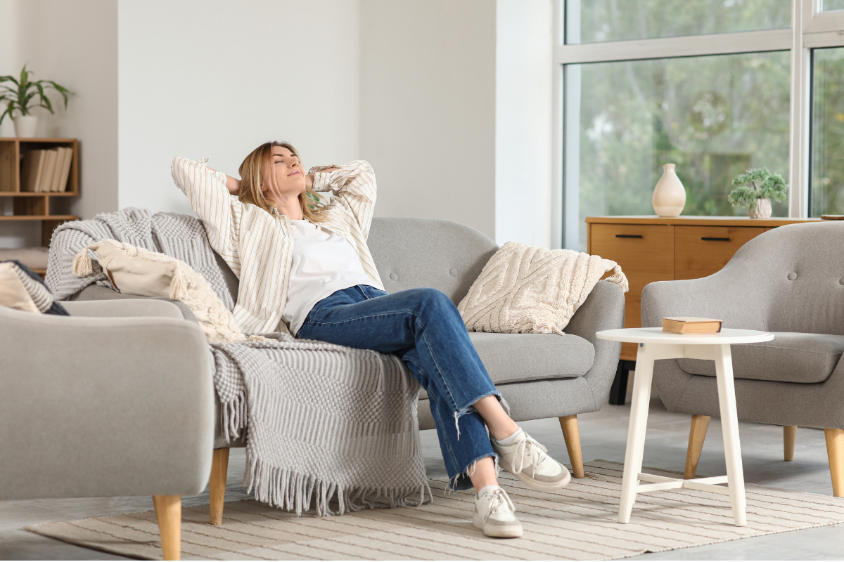 Woman relaxing on a grey sofa in a bright, sunlit living room.