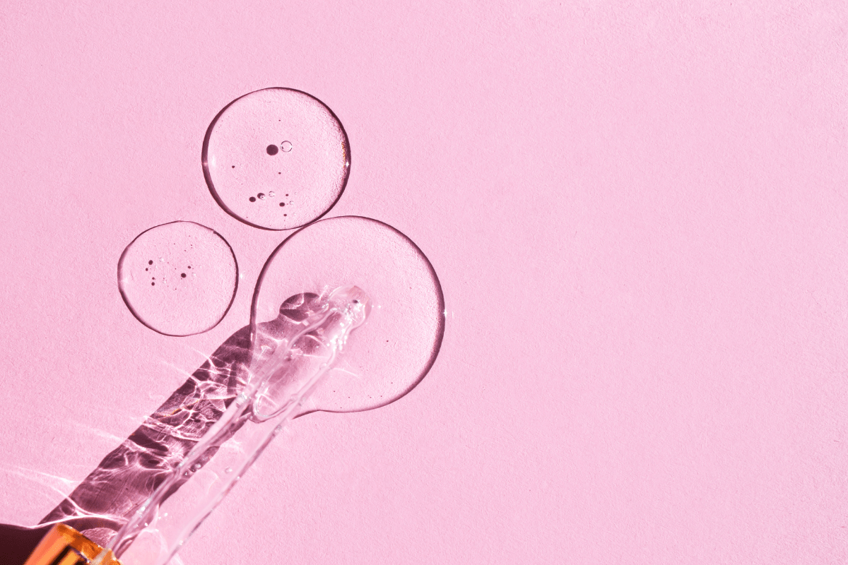 A dropper with clear serum and liquid droplets on a pink background.