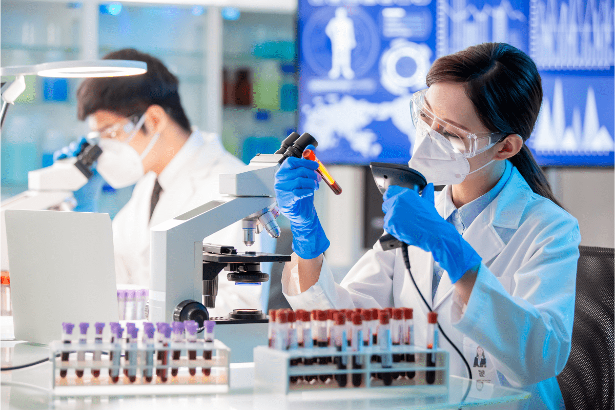 Two lab technicians wearing protective masks and glasses, analysing blood samples in a laboratory.