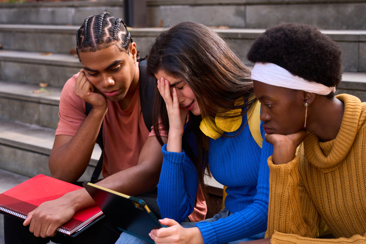 Three students looking stressed while sitting on steps with a laptop.