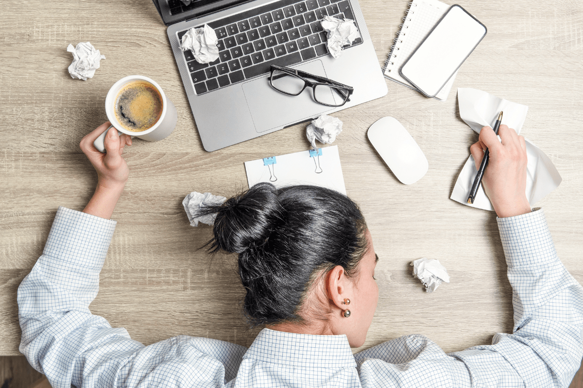 Exhausted person with head on desk surrounded by crumpled papers, laptop, and coffee cup.