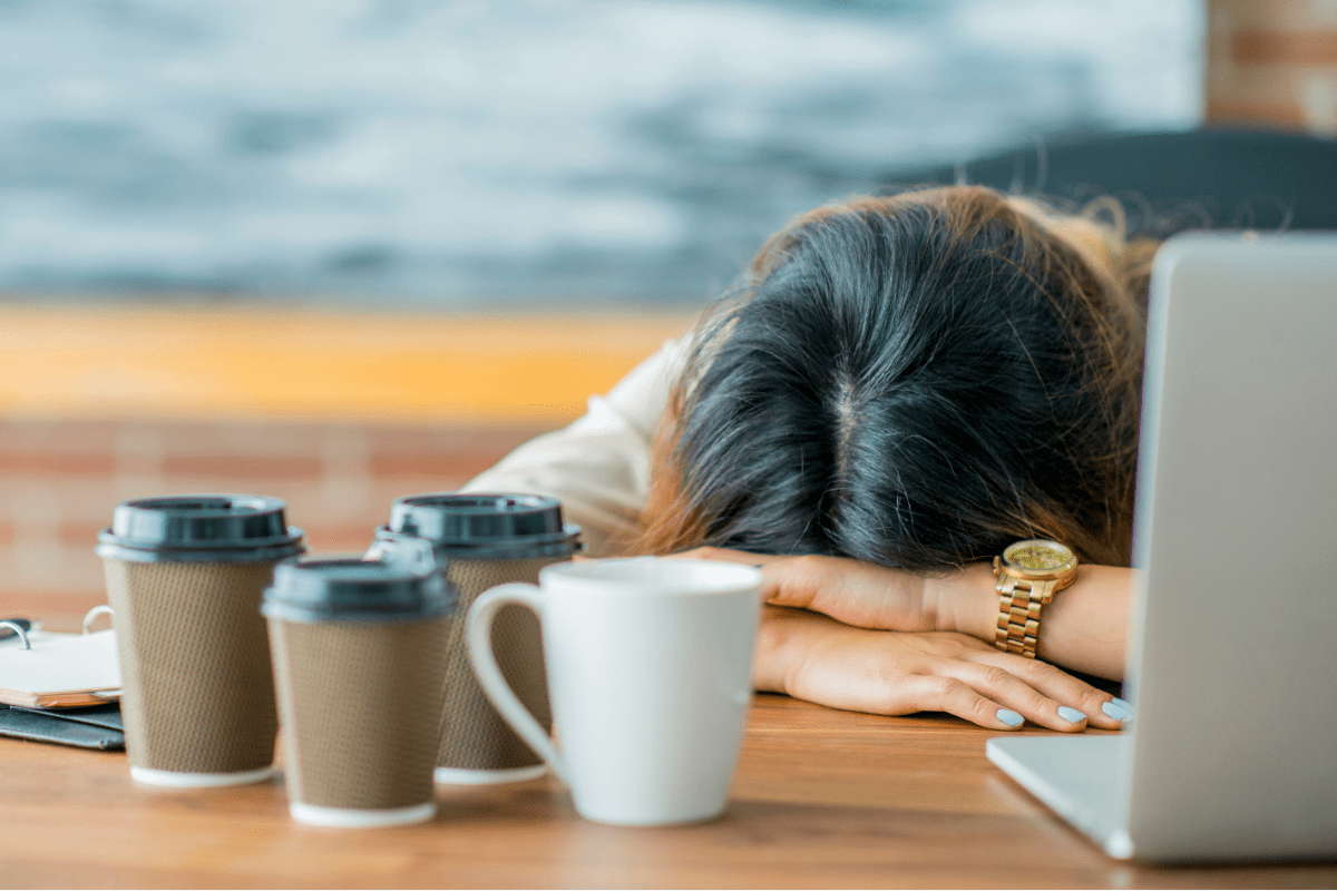 Exhausted woman with head on desk surrounded by coffee cups.