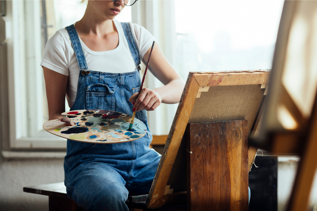 Person wearing overalls painting on a canvas, holding a paintbrush and a palette with various colours in a bright, naturally lit studio.