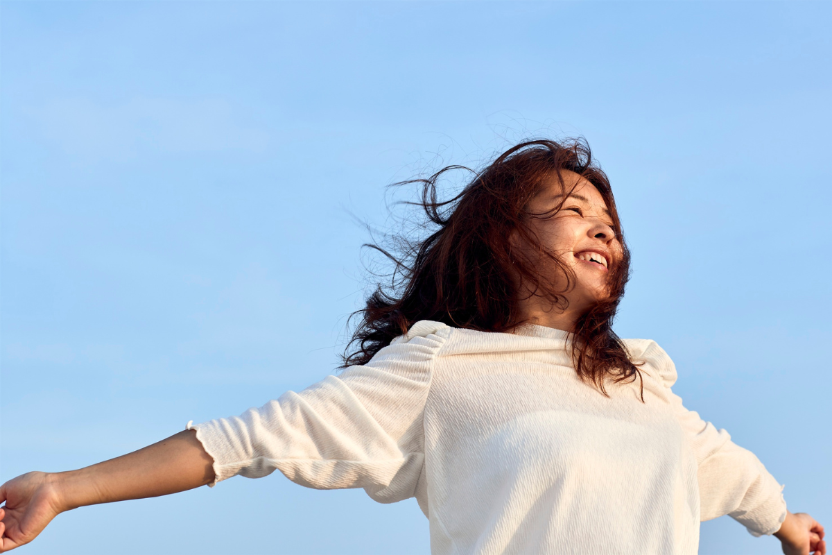 Smiling woman with arms outstretched and windblown hair beneath a blue sky.