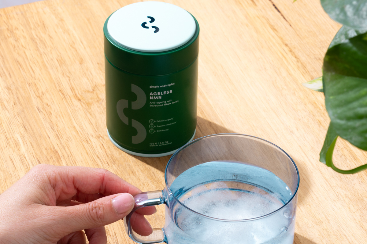 Hand mixing a glass of water next to a container of Ageless NMN on a wooden table.