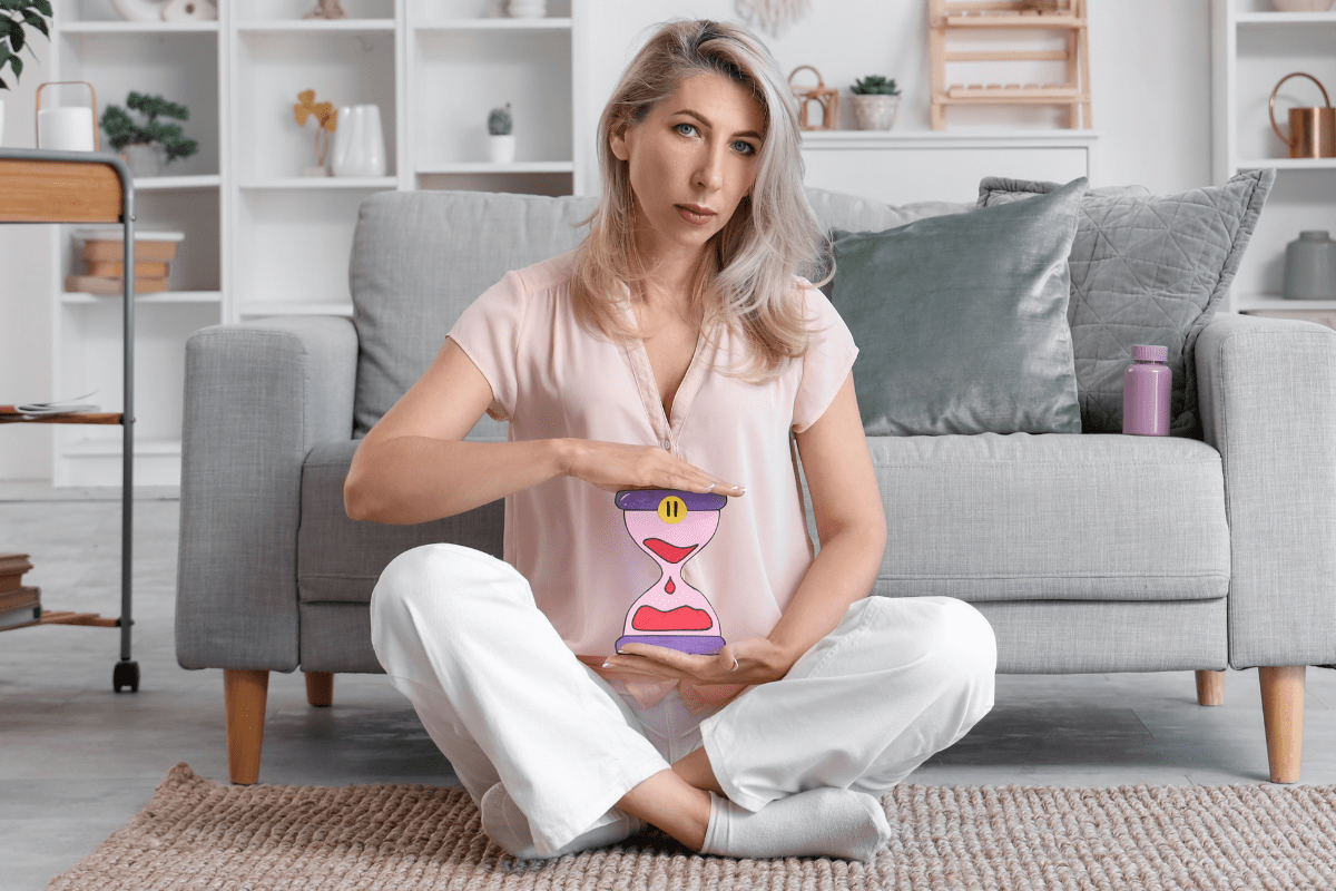 Woman sitting cross-legged indoors, holding a cartoon hourglass illustration over her abdomen.
