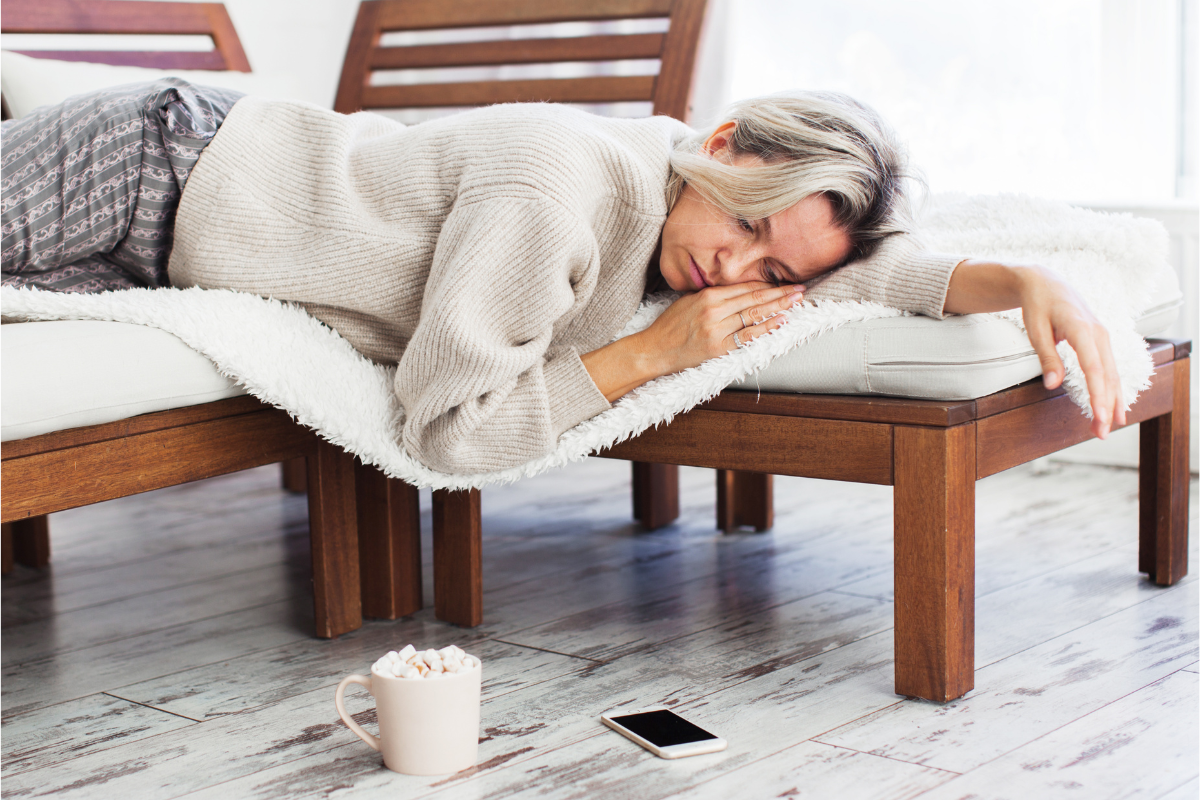 Woman lying on a bench looking tired, with a mug and phone on the floor nearby.