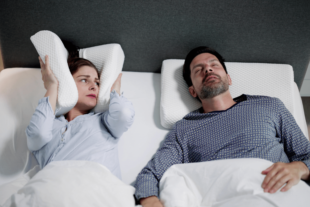 Woman covers her ears with a pillow while a man snores beside her in bed.