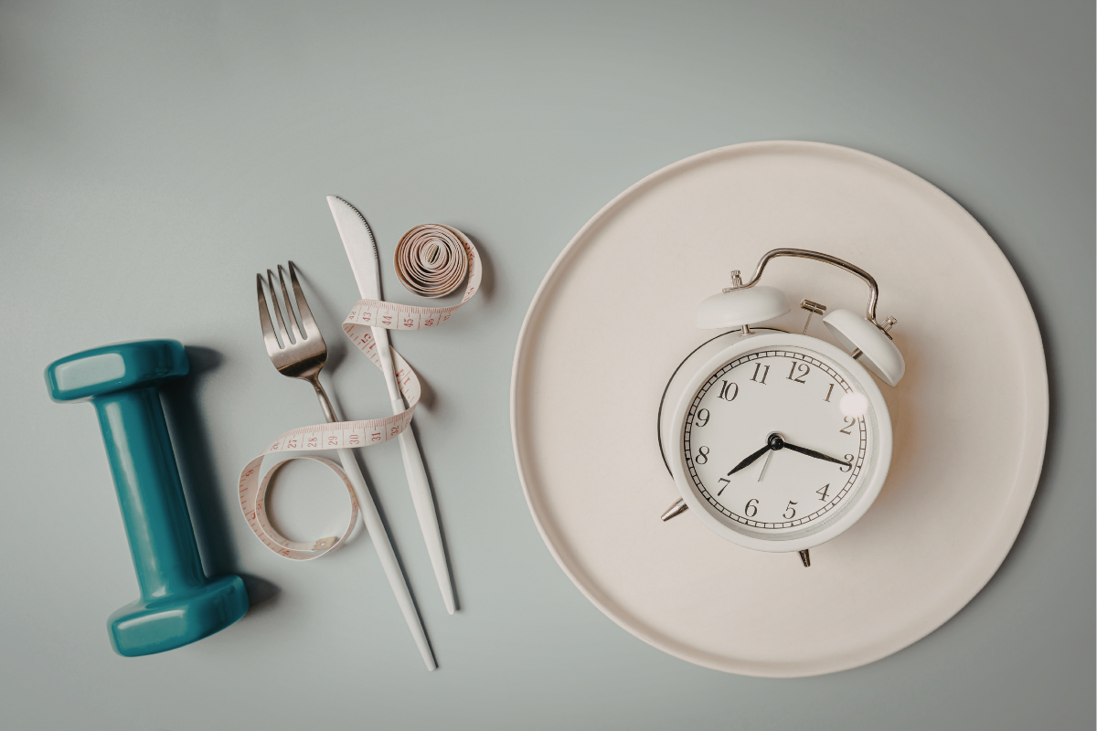 Plate with alarm clock, fork and knife, measuring tape, and dumbbell symbolising fasting and metabolism.