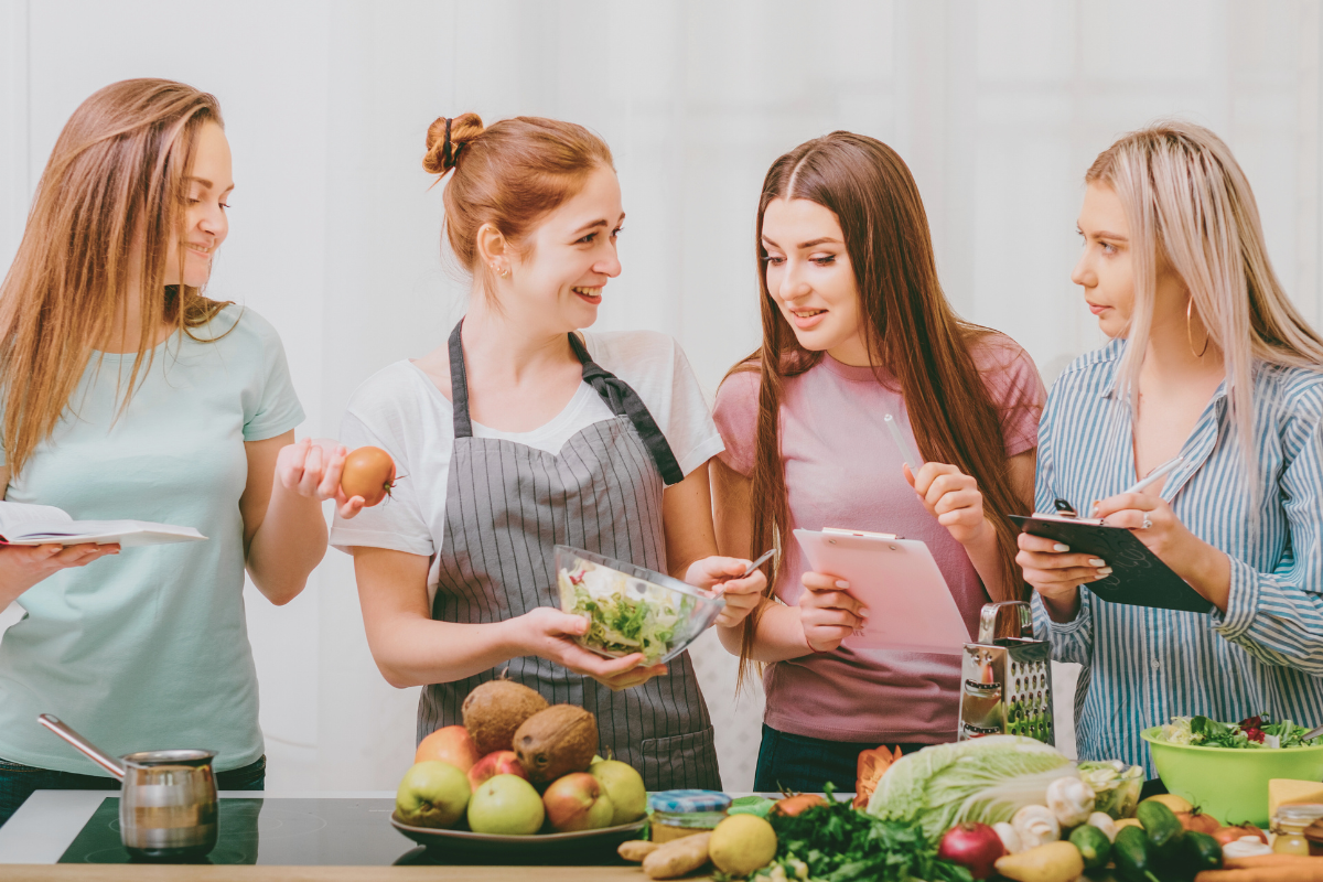 Four women preparing healthy food together in a bright kitchen, smiling and holding fresh ingredients and notebooks.