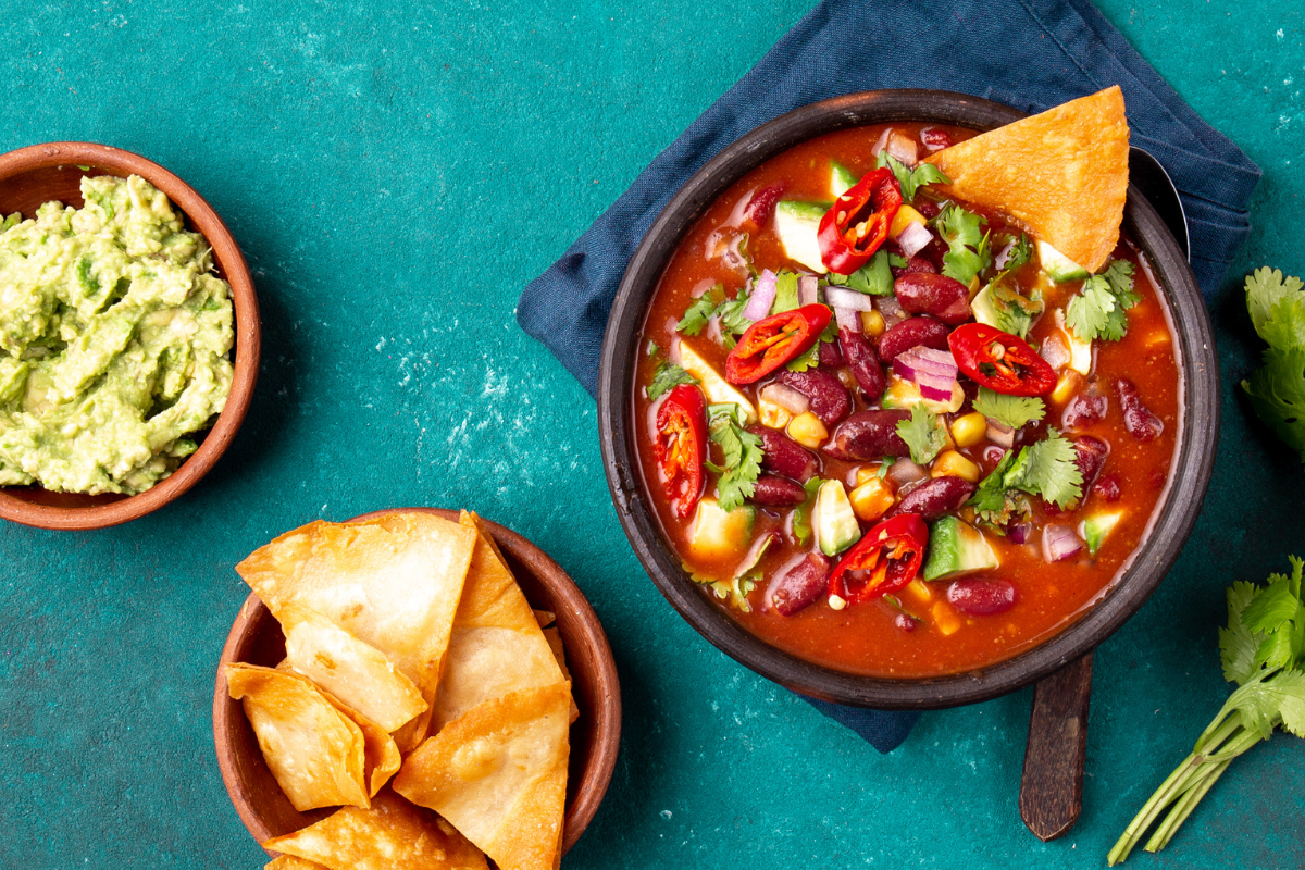 A bowl of spicy black bean chilli topped with fresh vegetables and herbs, served with homemade nachos and a side of guacamole.