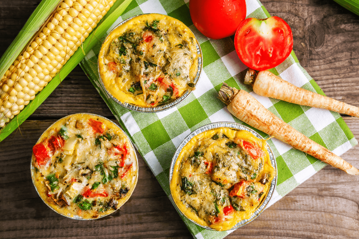 Three baked egg muffins in foil cups, filled with vegetables and herbs, placed on a green cloth alongside fresh corn, tomatoes, and parsnips.