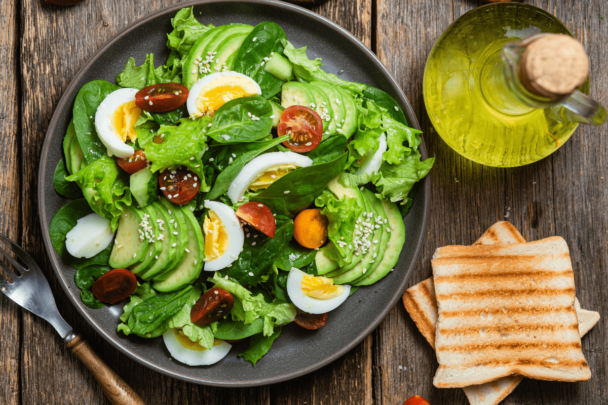 Fresh spinach salad with boiled eggs, avocado, cherry tomatoes, and sesame seeds, served with toasted bread and olive oil on a wooden table.