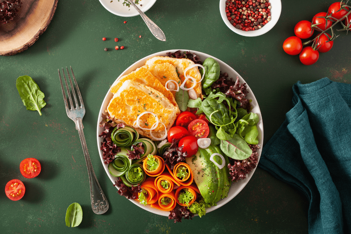 Colourful salad bowl with grilled halloumi, fresh greens, cherry tomatoes, cucumber, avocado, and carrot ribbons.