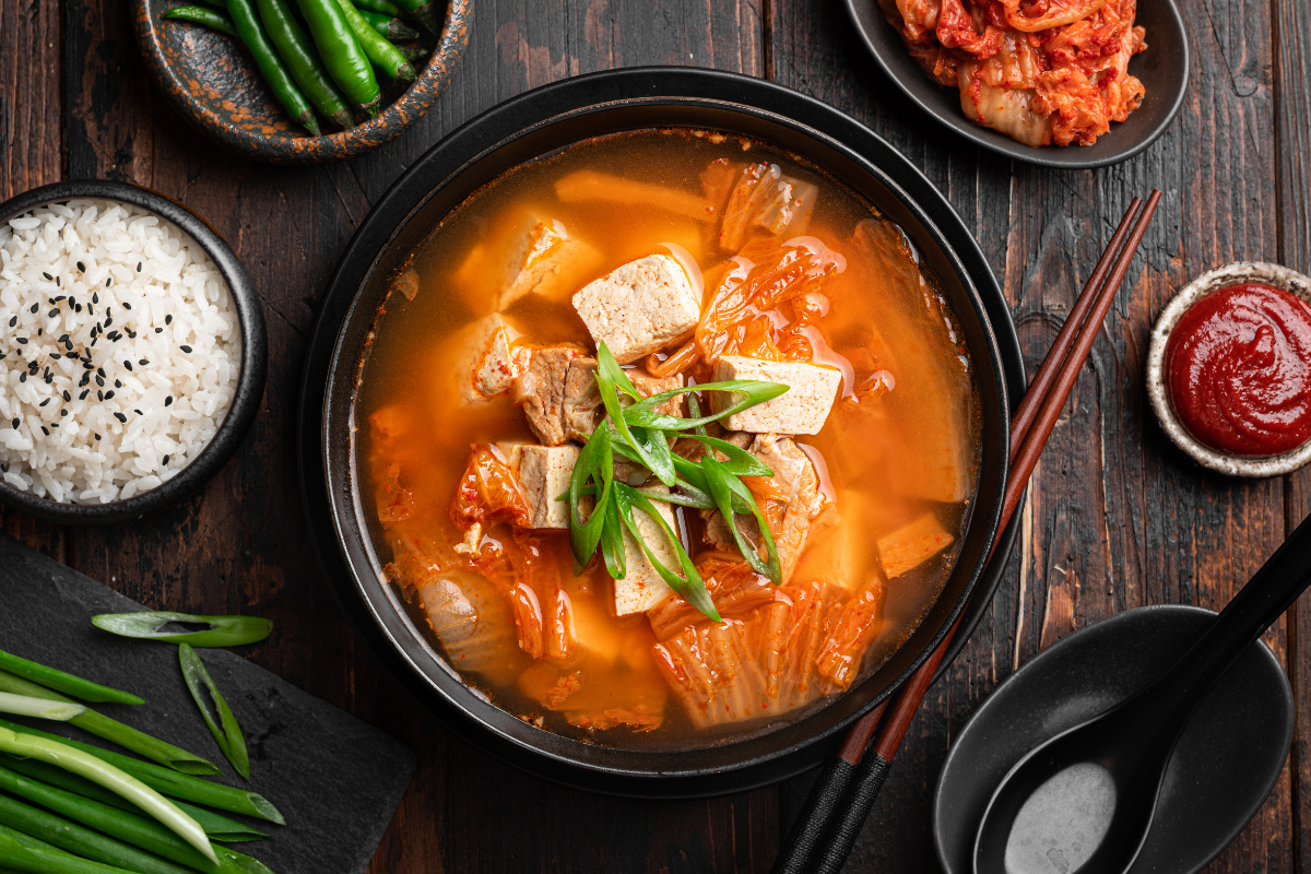 Bowl of kimchi soup with tofu and green onions, served with rice and side dishes.