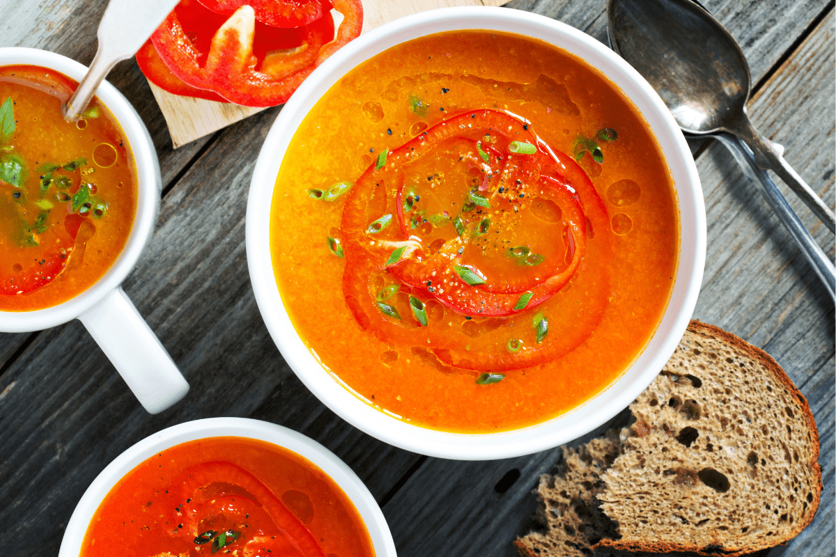 A bowl of vibrant roasted red pepper soup garnished with fresh herbs and served with rustic bread.