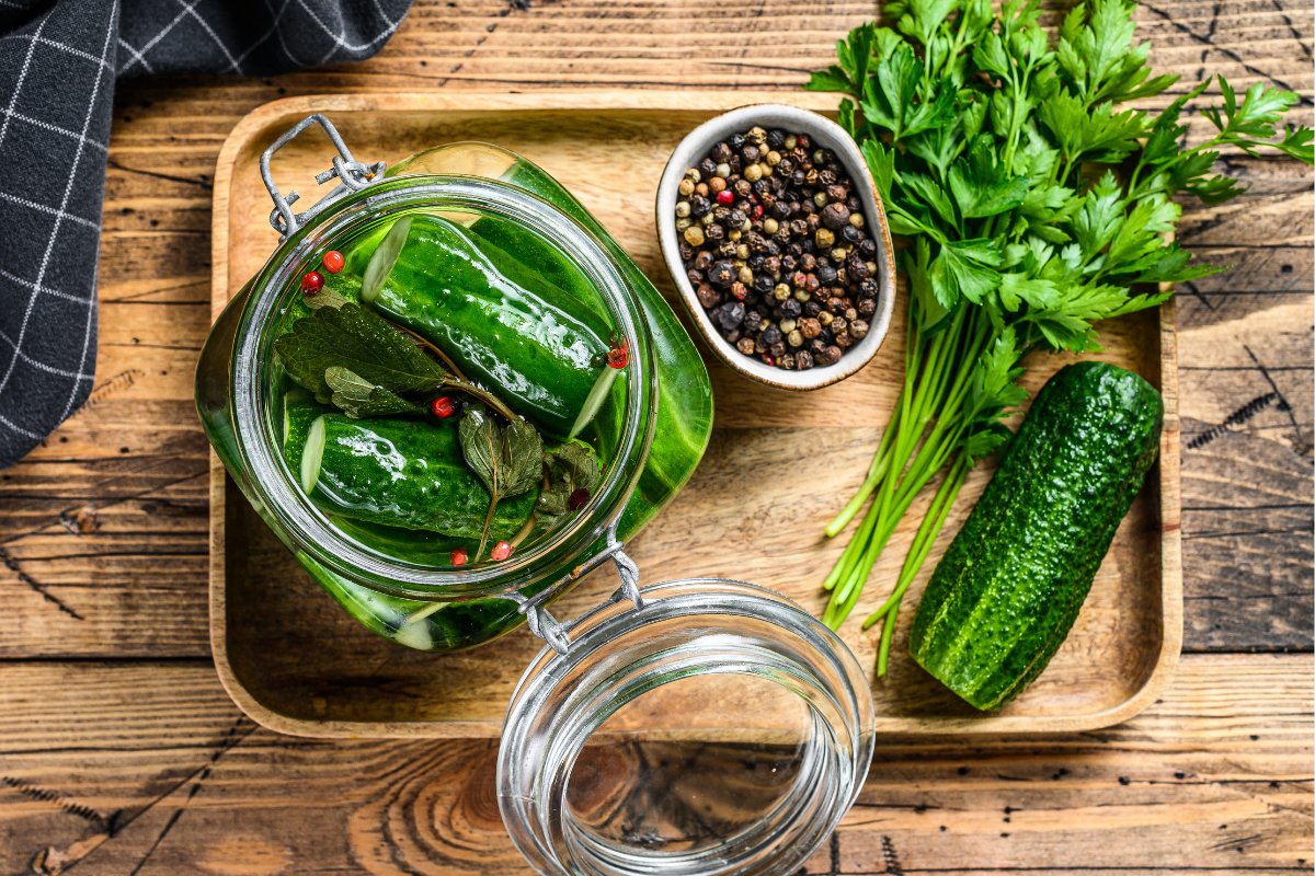 Jar of pickled cucumbers with herbs and peppercorns on a wooden tray.
