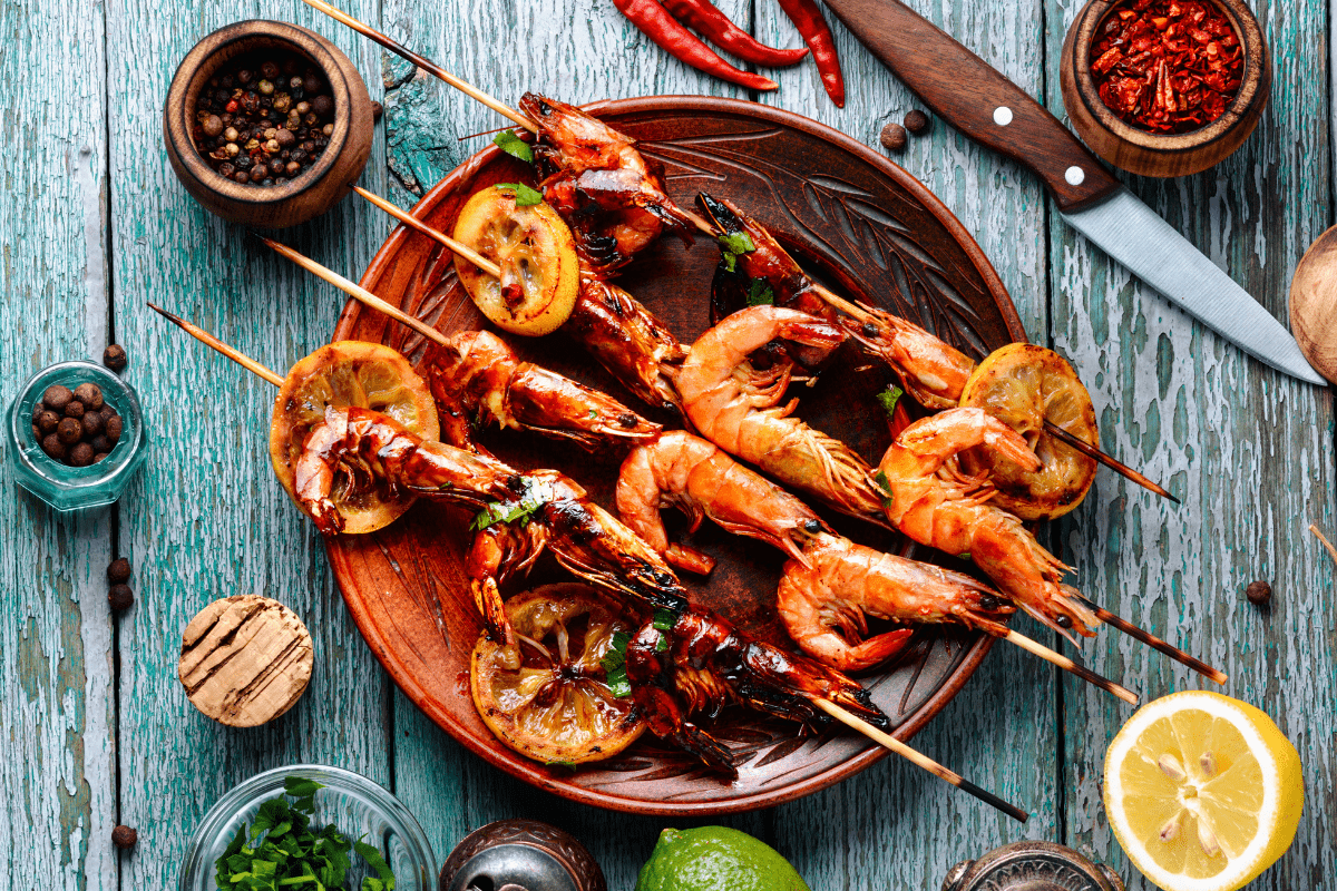 Grilled shrimp skewers with lemon slices served on a wooden plate, surrounded by fresh herbs and spices.