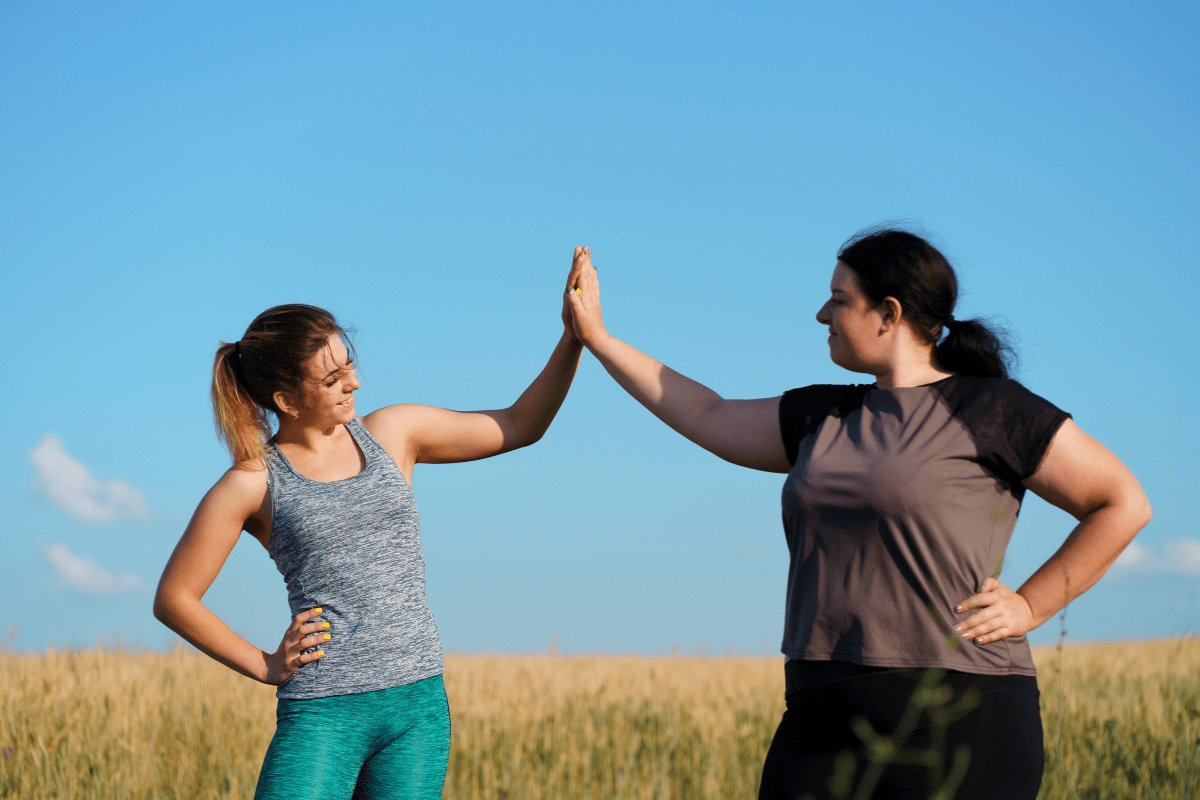 Two women standing outdoors, one woman is slim, and the other is curvier, both in activewear against a bright blue sky.