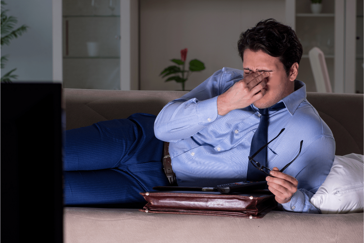 A man in a blue shirt and tie sits on a couch, rubbing his eyes.