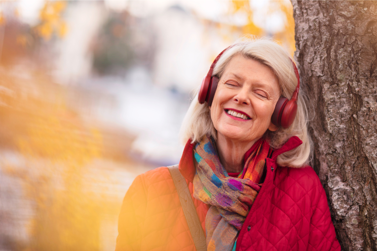 An elderly woman leans against a tree with a smile on her face, enjoying music through her red headphones.